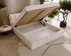 A white upholstered bed with its storage compartment open, revealing the slatted base and interior. The bed is in a room with wooden flooring and a rug. Produktbild-Vorschau