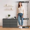 A woman stands next to a dresser, with a height measurement scale on the right side of the image. Produktbild-Vorschau