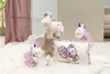 A collection of plush unicorn toys and a unicorn-themed pillow arranged on a fluffy white rug, with a draped pink curtain in the background. Produktbild-Vorschau