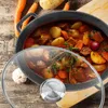 A close-up view of a pot of stew with ingredients like carrots and potatoes around it, placed on a wooden surface. Náhled obrázku produktu