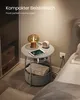 A small, round side table with a lamp, tablet, and storage basket next to a bed. Produktbild-Vorschau