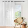 A white sheer curtain is partially drawn across a window, with a wicker chair and a small table with flowers next to it. Produktbild-Vorschau