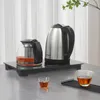 A glass teapot and a stainless steel electric kettle are displayed on a black heating tray, with two small glasses of tea in the foreground. Náhled obrázku produktu