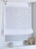 A white sheer Roman blind with embroidered floral pattern hanging in a window frame. Decorative heart ornaments and a small stone sculpture are on the windowsill below. Produktbild-Vorschau