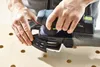 Hands holding a Festool Akku-Deltaschleifer DTSC 400 3,0 I-Plus over a wooden surface with holes. Voir les images du produit