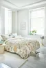 A bedroom with a bed dressed in floral bedding, natural light streaming in from large windows. Produktbild-Vorschau