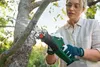 A woman is using a cordless reciprocating saw to cut a tree branch outdoors. Produktbild-Vorschau