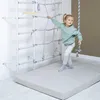 A young girl is playing on a gymnastics wall with a rope and a wooden bar, on a padded mat. Produktbild-Vorschau