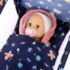 A close-up view of a doll lying in a travel crib with a pink hood and a navy blue blanket with a floral and butterfly pattern. Produktbild-Vorschau
