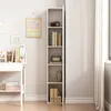 A tall, narrow white bookshelf is shown standing against a light gray wall, next to a white desk with some items on it. Podgląd zdjęcia produktu
