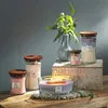 A collection of Woodwick candles with wooden lids, arranged on a wooden surface with a vase of greenery in the background. Náhled obrázku produktu
