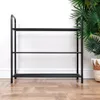A black metal shoe rack with three shelves against a white wall and a wooden floor. Produktbild-Vorschau