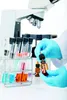 A person in a lab coat and blue gloves is holding a dropper over a small brown bottle, with test tubes and a microscope in the background. Produktbild-Vorschau