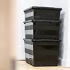 Three black storage boxes stacked on a light wooden floor next to a white wall. Anteprima dell’immagine prodotto