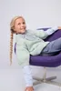 A young girl with blonde pigtails sits in a purple chair, looking up and to the side. She is wearing a light green knitted sweater over a light blue collared shirt and gray pants. Produktbild-Vorschau