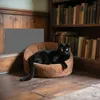 A black cat is resting in a brown cat bed in front of a bookshelf filled with books. A gray textured mat is on the wall behind the cat bed. Produktbild-Vorschau
