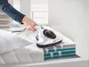 A hand is using a white handheld vacuum cleaner on a mattress, with a cutaway showing airflow within the mattress layers. Produktbild-Vorschau