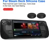 A black silicone case for a Steam Deck gaming console is shown, along with two black joystick caps. The console screen displays game art. Produktbild-Vorschau