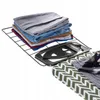 A stack of folded clothes rests on a black wire rack, next to a black iron rest on a green and white chevron-patterned ironing board. Produktbild-Vorschau