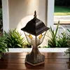 An outdoor lamp is placed on a wooden table in front of plants and a wall. Produktbild-Vorschau