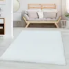 A white shaggy rug is laid out on a light wooden floor in a living room setting. A modern wooden sofa with beige and white cushions is visible in the background, along with a small white side table and a plant. Anteprima dell’immagine prodotto