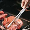 A hand is using kitchen tongs to flip a piece of meat on a grill. Produktbild-Vorschau