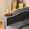 A close-up of a dark gray sofa with a built-in cup holder containing a glass of orange liquid. A guitar is visible in the background. Produktbild-Vorschau