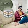 A woman is sitting on a couch, reading a book and wrapped in a burgundy heated blanket. Produktbild-Vorschau