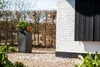 A tall, rectangular planter with plants sits on a paved patio next to a white brick building with black shutters. Produktbild-Vorschau
