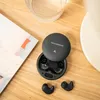A pair of black wireless earbuds and their charging case are arranged on a wooden surface next to a magazine and a glass of water. Produktbild-Vorschau