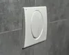 A close-up shot of a white toilet flush plate mounted on a gray tiled wall. Náhled obrázku produktu