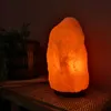 A Himalayan salt lamp is lit on a wooden shelf next to a potted plant. Produktbild-Vorschau