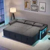 A modern L-shaped sofa bed with integrated LED lighting and USB ports, set up in a living room environment with artwork on the wall and a city view through the window. Produktbild-Vorschau