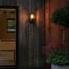 An outdoor wall light illuminates a wooden wall next to a glass door and a potted plant. Produktbild-Vorschau