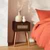 A dark wood nightstand with a rattan drawer front sits next to a bed with orange bedding. A vase with dried flowers and a small white decorative object are on top of the nightstand. Produktbild-Vorschau