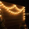 String lights are hung on a wooden wall at night, with a lantern and some greenery in the foreground. Produktbild-Vorschau