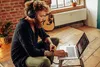 A man with headphones is sitting on the floor in front of a laptop, working on music production. An acoustic guitar is visible in the background. Produktbild-Vorschau
