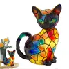 A decorative stained-glass style cat figurine with colorful panels, shown on a white background. A smaller version of the same figurine is visible in the lower left corner, next to a vase of flowers. Produktbild-Vorschau