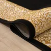 A close-up view of a rolled-up rug with a black border and a cream and gold patterned design. Produktbild-Vorschau