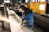 A worker in a factory is using an angle grinder on a large metal beam, creating sparks. Produktbild-Vorschau