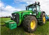 A green tractor is parked in a grassy field under a blue sky with white clouds. Other tractors are visible in the background. Produktbild-Vorschau