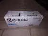A cardboard box for a Kyocera toner cartridge sits on a brick surface. Produktbild-Vorschau