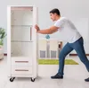A man is pushing a white cabinet with glass doors in a room with dining furniture. Produktbild-Vorschau