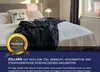 A bed with a dark blanket and decorative pillows, with a promotional seal and text overlay. Produktbild-Vorschau