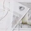 Close-up of a beige roller blind with a decorative embroidered pattern and a silver eyelet, placed over a magazine or book with text and images of milk bottles. Produktbild-Vorschau