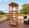 A wooden bird aviary stands on a tiled patio, with a railing and trees in the background. Produktbild-Vorschau