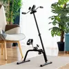 A black pedal exerciser is placed on a rug in front of a chair and plants. Produktbild-Vorschau