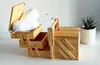 A wooden sewing box with open compartments containing balls of white yarn and knitting needles, placed on a white surface next to a potted plant. Podgląd zdjęcia produktu