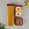 Three food storage containers with red lids are arranged on a light gray surface. The largest container on the left is filled with spaghetti. The middle container holds dried apple rings, and the smallest container at the bottom contains crackers. Anteprima dell’immagine prodotto