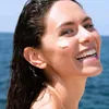 A woman is smiling while applying sunscreen to her face outdoors with the ocean in the background. Náhled obrázku produktu